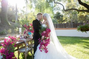 Rancho las lomas outdoor wedding shoot bride strapless tulle ballgown with beaded bodice and sweetheart neckline with long lace detail veil and groom black notch lapel tuxedo with white dress shirt and black bow tie with bright pink floral boutonniere bride holding bright pink floral bridal bouquet standing by table