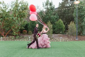 Los angeles valentine theme wedding shoot at the gardens at los robles bride pink ballgown dress with ruffled skirt and dark pink and white floral bridal bouquet with groom burgundy tuxedo with black dress shirt and black bow tie with red floral boutonniere walking and holding balloons