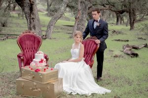 Valentine styled wedding shoot bride chiffon gown with halter straps and sweetheart neckline with groom navy blue suit with notch lapel with matching vest and white dress shirt with red bow tie and white floral boutonniere sitting in lounge furniture