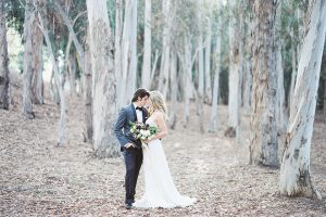 Into the woods outdoor romantic wedding shoot bride chiffon gown with thin spahetti straps and strap design on back with lace detail and sweetheart neckline with groom charcoal grey tuxedo coat with black lapels and black pants with white dress shirt and black bow tie hugging and bride holding white and purple floral bridal bouquet