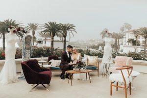 Dana point outdoor wedding shoot at monarch beach resort bride form fitting lace gown with thin straps and plunging neckline with groom burgundy tuxedo with matching vest and black bow tie with floral boutonniere sitting on couch