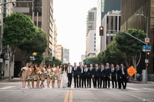 Downtown los angeles wedding bride form fitting lace gown with illusion back and high neckline with crystal hair piece with groom charcoal grey tuxedo with black shawl lapel and white dress shirt with long black skinny tie and white floral boutonniere with bridesmaids short gold sequined dresses and groomsmen black tuxedos with long black skinny ties