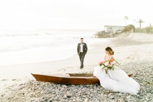 Orange county beach wedding at ole hanson beach club bride ball gown with thin straps and sweetheart neckline with low back design and lace details with groom charcoal grey tuxedo with white dress shirt and black bow tie bride sitting in boat with groom walking