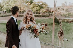 San diego outdoor wedding shoot at the san diego zoo and safari park bride white chiffon gown with lace sleeves and thin straps with plunging neckline and groom burgundy shawl lapel tuxedo with white dress shirt and long black skinny tie and black shoes standing and hugging with giraffe in background bride holding white and orange floral bridal bouquet