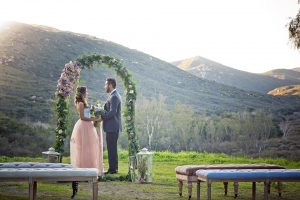 San diego outdoor woodland wedding shoot bride blush pink chiffon strapless gown with a sweetheart neckline and groom navy blue shawl lapel tuxedo with a matching vest and white dress shirt with a long pink striped tie and white floral boutonniere standing and holding hands during ceremony bride holding white floral bridal bouquet