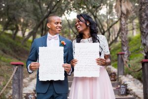 Rancho las lomas outdoor engagement shoot bride light pink tulle skirt with a white lace short sleeve top with groom slate blue notch lapel suit with a white dress shirt and blush pink bow tie with a pink floral boutonniere standing and holding vows during ceremony