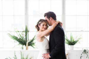 Downtown los angeles wedding shoot at fd photo studio bride form fitting simple gown with a high halter neckline and groom black notch lapel tuxedo by michael kors with a white dress shirt and black bow tie hugging and smiling with greenery decor