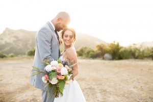 Los angeles outdoor wedding at brookview ranch bride two piece wedding dress with tulle skirt and crop top bodice with thin straps and a sweetheart neckline and groom heather grey notch lapel suit with a matching vest and white dress shirt with a long navy blue tie and white floral boutonniere hugging and bride holding white and pink floral bridal bouquet