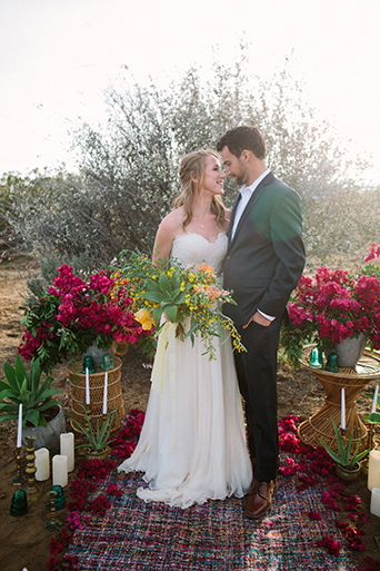 Anza valley rustic outdoor wedding at the alpaca farm bride strapless chiffon gown with a sweetheart necklace holding green and yellow floral bridal bouquet with groom navy blue suit with white dress shirt and no tie standing by pink floral arch for ceremony