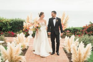 Dana point wedding at doheny house bride strapless a line gown with a beaded bodice and long veil with lace arm sleeves with groom black tuxedo with white dress shirt and black bow tie with orange floral boutonniere bride holding white and peach floral bridal bouquet walking down aisle holding hands