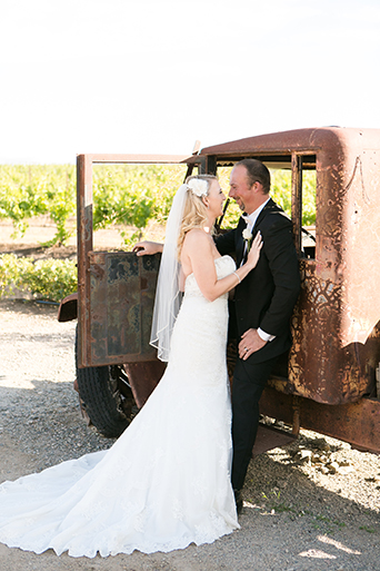 Temecula outdoor wedding at villa de amore vineyard bride strapless mermaid style gown with crystal belt and medium veil with groom black tuxedo with white dress shirt and white bow tie with matching vest and white floral boutonniere hugging by brown vintage truck
