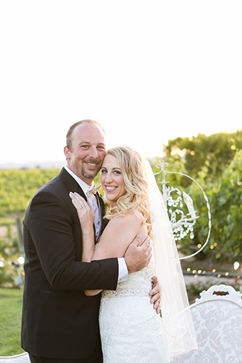 Temecula outdoor wedding at villa de amore vineyard bride strapless mermaid style gown with crystal belt and medium veil with groom black tuxedo with white dress shirt and white bow tie with matching vest and white floral boutonniere standing by white lounge furniture decor