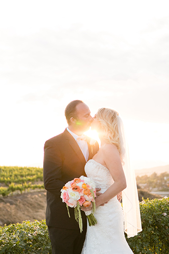 Temecula outdoor wedding at villa de amore vineyard bride strapless mermaid style gown with crystal belt and medium veil with groom black tuxedo with white dress shirt and white bow tie with matching vest and white floral boutonniere kissing at sunset
