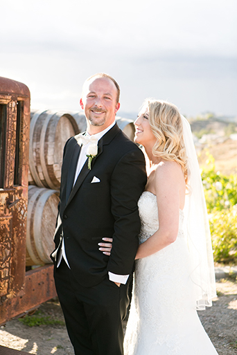 Temecula outdoor wedding at villa de amore vineyard bride strapless mermaid style gown with crystal belt and medium veil with groom black tuxedo with white dress shirt and white bow tie with matching vest and white floral boutonniere standing by brown vintage truck