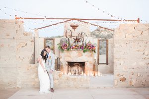 Moroccan inspired outdoor wedding shoot at the ruin venue bride lace form fitting gown with plunging neckline and beaded short sleeves with crystal headpiece and groom light grey suit with matching vest and white dress shirt with long blue tie and bright orange floral boutonniere sitting by ceremony set up with white candle decor and hanging lights