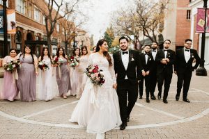 Orange county wedding at the estate on second bride lace ball gown with long sleeves and high neckline with crystal hair piece and groom black tuxedo with white dress shirt and black bow tie with white pocket square with bridesmaids mix and matched dresses with groomsmen black tuxedos with white shirts and black bow ties walking bride holding pink and red floral bridal bouquet