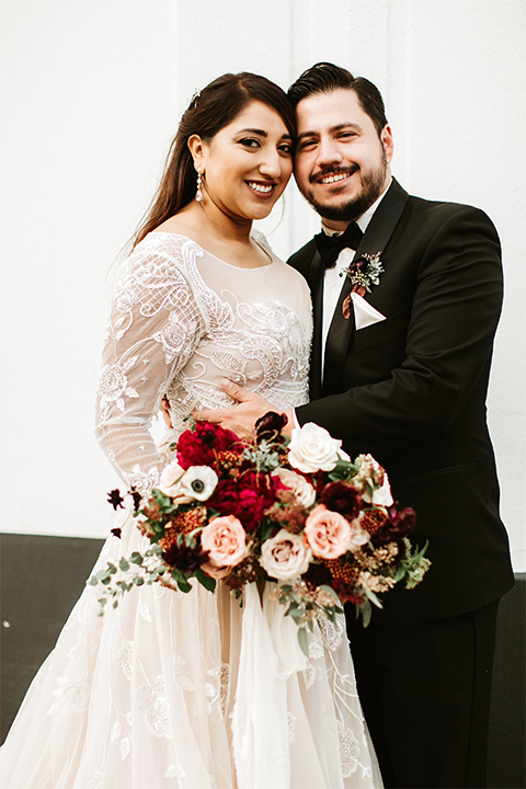 Orange county wedding at the estate on second bride lace ball gown with long sleeves and high neckline with crystal hair piece and groom black tuxedo with white dress shirt and black bow tie with white pocket square hugging bride holding pink and red floral bridal bouquet