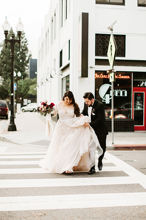 Orange county wedding at the estate on second bride lace ball gown with long sleeves and high neckline with crystal hair piece and groom black tuxedo with white dress shirt and black bow tie with white pocket square walking across the street bride holding pink and red floral bridal bouquet