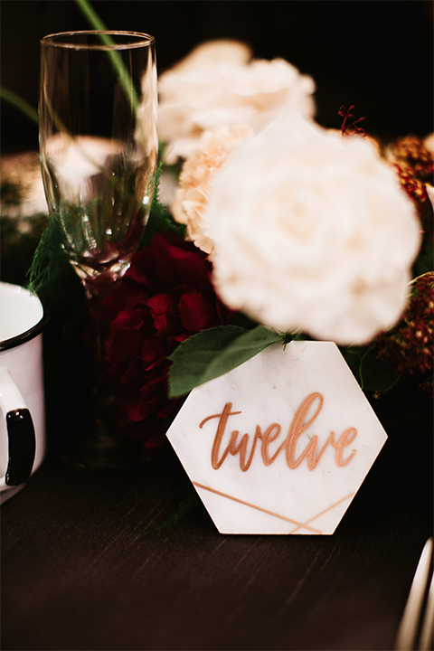 Orange county wedding at the estate on second dark brown wood table with gold place settings and white and green flower centerpiece decor with white glasses and candles and black chairs and white marble table numbers with gold calligraphy writing