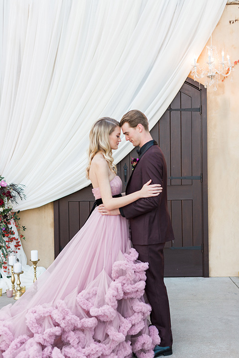 Los angeles valentine theme wedding shoot at the gardens at los robles bride pink ballgown dress with ruffled skirt and dark pink and white floral bridal bouquet with groom burgundy tuxedo with black dress shirt and black bow tie with red floral boutonniere hugging