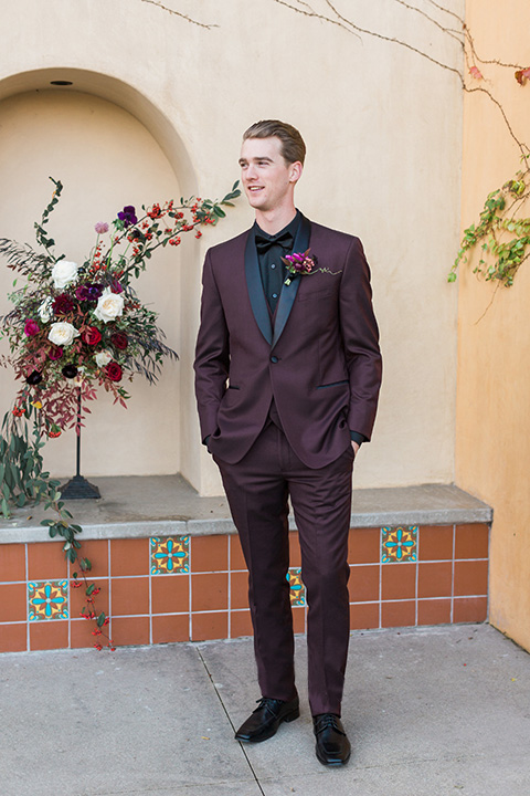 Los angeles valentine theme wedding shoot at the gardens at los robles groom burgundy tuxedo with black dress shirt and black bow tie with red floral boutonniere standing with hands in pockets