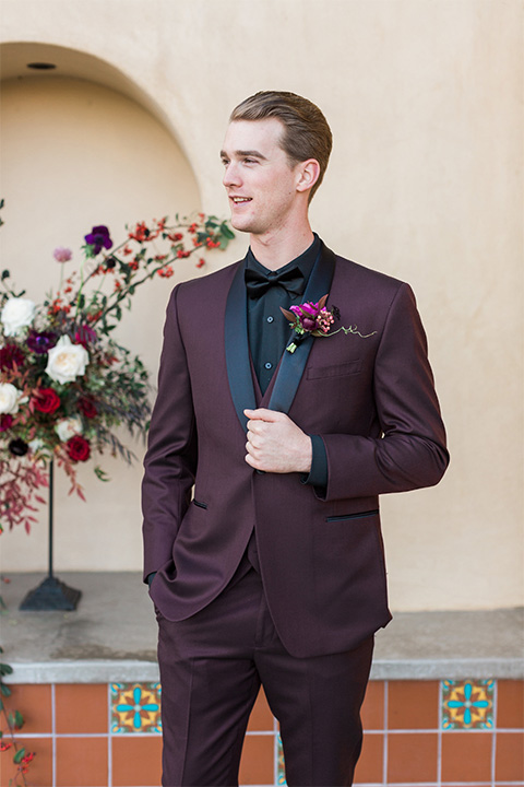 Los angeles valentine theme wedding shoot at the gardens at los robles groom burgundy tuxedo with black dress shirt and black bow tie with red floral boutonniere standing with hands in pockets