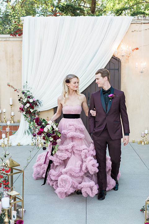 Los angeles valentine theme wedding shoot at the gardens at los robles bride pink ballgown dress with ruffled skirt and dark pink and white floral bridal bouquet with groom burgundy tuxedo with black dress shirt and black bow tie with red floral boutonniere walking and holding hands