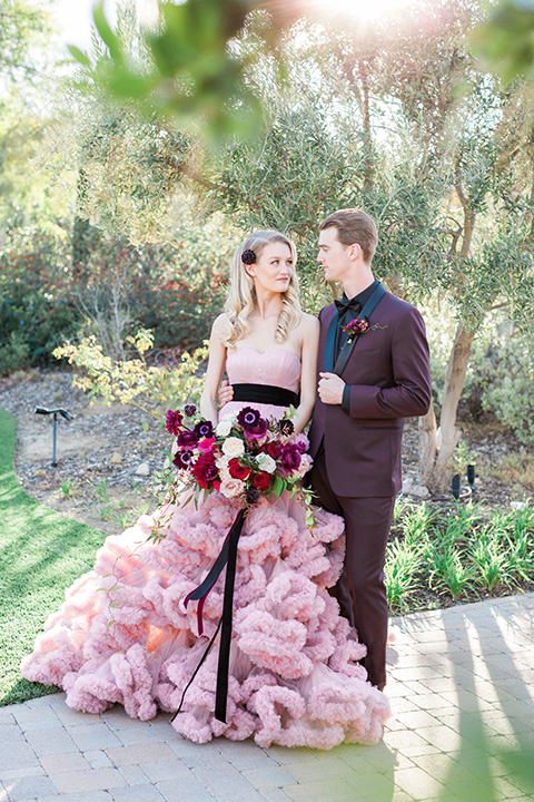 Los angeles valentine theme wedding shoot at the gardens at los robles bride pink ballgown dress with ruffled skirt and dark pink and white floral bridal bouquet with groom burgundy tuxedo with black dress shirt and black bow tie with red floral boutonniere walking and holding hands