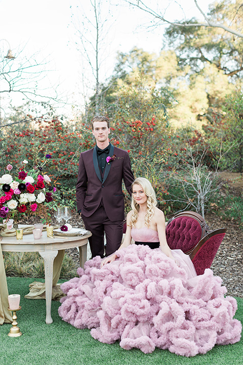 Los angeles valentine theme wedding shoot at the gardens at los robles bride pink ballgown dress with ruffled skirt and dark pink and white floral bridal bouquet with groom burgundy tuxedo with black dress shirt and black bow tie with red floral boutonniere sitting in vintage lounge furniture