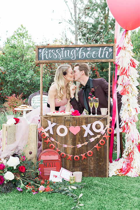 Los angeles valentine theme wedding shoot at the gardens at los robles bride pink ballgown dress with ruffled skirt and dark pink and white floral bridal bouquet with groom burgundy tuxedo with black dress shirt and black bow tie with red floral boutonniere kissing in kissing booth