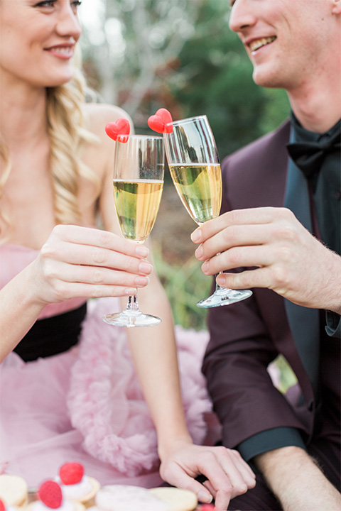 Los angeles valentine theme wedding shoot at the gardens at los robles bride pink ballgown dress with ruffled skirt and dark pink and white floral bridal bouquet with groom burgundy tuxedo with black dress shirt and black bow tie with red floral boutonniere holding champagne glasses