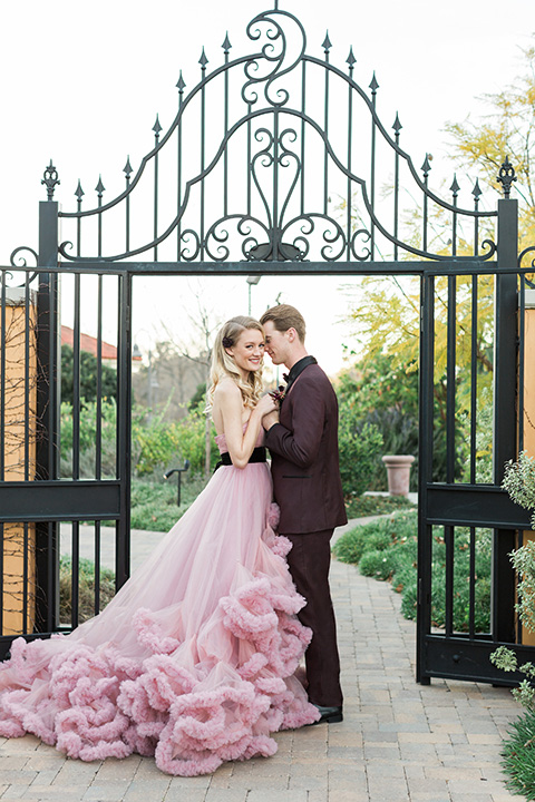 Los angeles valentine theme wedding shoot at the gardens at los robles bride pink ballgown dress with ruffled skirt and dark pink and white floral bridal bouquet with groom burgundy tuxedo with black dress shirt and black bow tie with red floral boutonniere hugging