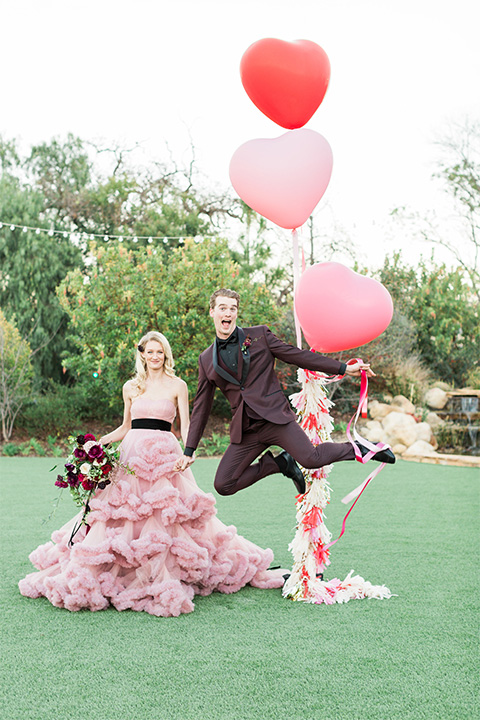 Los angeles valentine theme wedding shoot at the gardens at los robles bride pink ballgown dress with ruffled skirt and dark pink and white floral bridal bouquet with groom burgundy tuxedo with black dress shirt and black bow tie with red floral boutonniere holding balloons and groom jumping