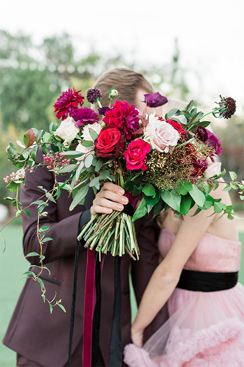 Los angeles valentine theme wedding shoot at the gardens at los robles bride pink ballgown dress with ruffled skirt and dark pink and white floral bridal bouquet with groom burgundy tuxedo with black dress shirt and black bow tie with red floral boutonniere kissing behind brides bouquet