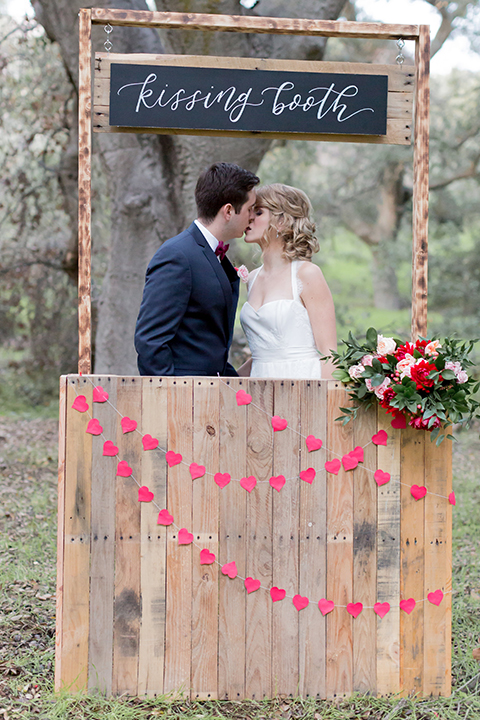 Valentine styled wedding shoot bride chiffon gown with halter straps and sweetheart neckline with groom navy blue suit with notch lapel with matching vest and white dress shirt with red bow tie and white floral boutonniere kissing in kissing booth