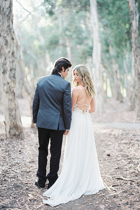 Into the woods outdoor romantic wedding shoot bride chiffon gown with thin spahetti straps and strap design on back with lace detail and sweetheart neckline with groom charcoal grey tuxedo coat with black lapels and black pants with white dress shirt and black bow tie holding hands