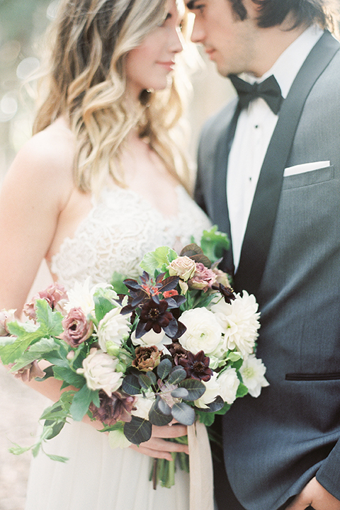 Into the woods outdoor romantic wedding shoot bride chiffon gown with thin spahetti straps and strap design on back with lace detail and sweetheart neckline with groom charcoal grey tuxedo coat with black lapels and black pants with white dress shirt and black bow tie bride holding white and purple floral bridal bouquet