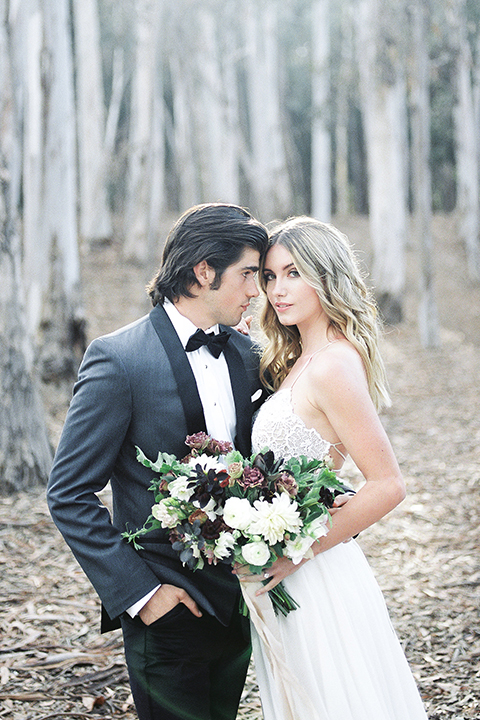 Into the woods outdoor romantic wedding shoot bride chiffon gown with thin spahetti straps and strap design on back with lace detail and sweetheart neckline with groom charcoal grey tuxedo coat with black lapels and black pants with white dress shirt and black bow tie bride holding white and purple floral bridal bouquet