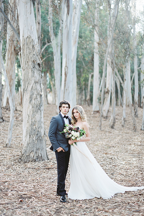 Into the woods outdoor romantic wedding shoot bride chiffon gown with thin spahetti straps and strap design on back with lace detail and sweetheart neckline with groom charcoal grey tuxedo coat with black lapels and black pants with white dress shirt and black bow tie bride holding white and purple floral bridal bouquet