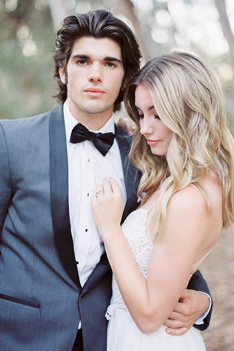 Into the woods outdoor romantic wedding shoot bride chiffon gown with thin spahetti straps and strap design on back with lace detail and sweetheart neckline with groom charcoal grey tuxedo coat with black lapels and black pants with white dress shirt and black bow tie hugging