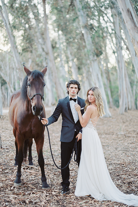 Into the woods outdoor romantic wedding shoot bride chiffon gown with thin spahetti straps and strap design on back with lace detail and sweetheart neckline with groom charcoal grey tuxedo coat with black lapels and black pants with white dress shirt and black bow tie with horse