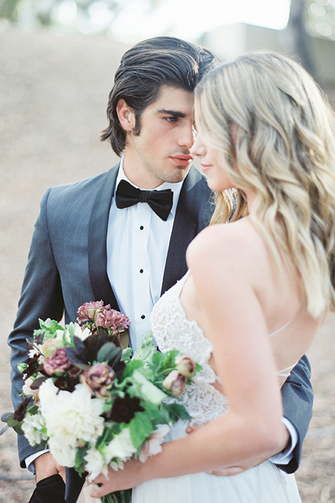 Into the woods outdoor romantic wedding shoot bride chiffon gown with thin spahetti straps and strap design on back with lace detail and sweetheart neckline with groom charcoal grey tuxedo coat with black lapels and black pants with white dress shirt and black bow tie hugging and bride holding white and purple floral bridal bouquet