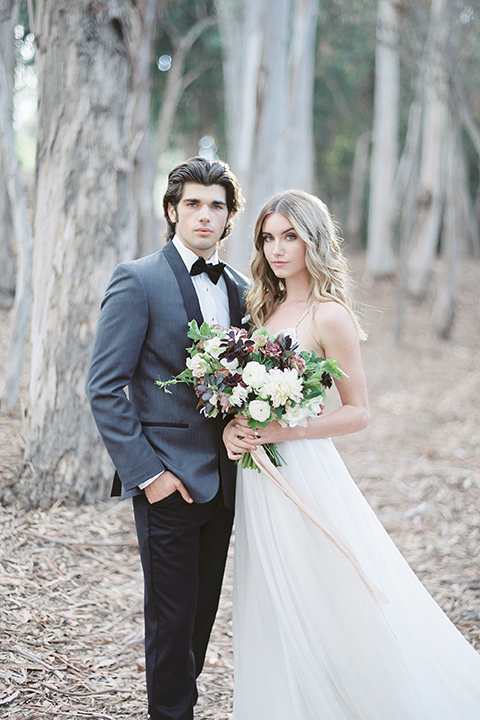 Into the woods outdoor romantic wedding shoot bride chiffon gown with thin spahetti straps and strap design on back with lace detail and sweetheart neckline with groom charcoal grey tuxedo coat with black lapels and black pants with white dress shirt and black bow tie bride holding white and purple floral bridal bouquet