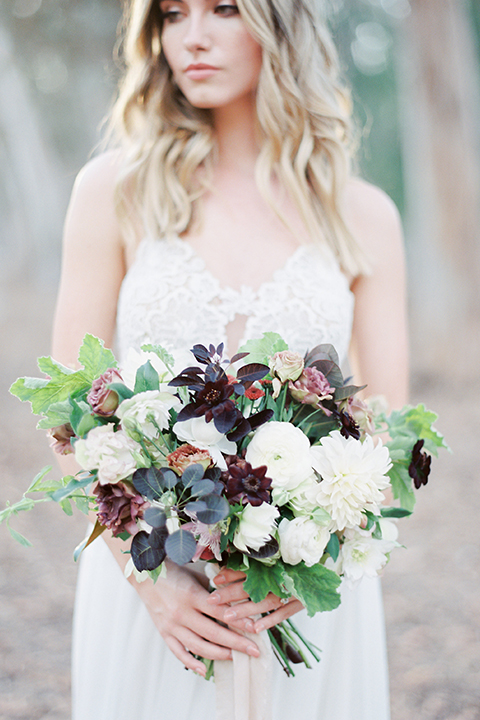 Into the woods outdoor romantic wedding shoot bride chiffon gown with thin spahetti straps and strap design on back with lace detail and sweetheart neckline holding white and purple floral bridal bouquet
