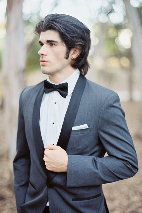 Into the woods outdoor romantic wedding shoot groom charcoal grey tuxedo with black lapels and black pants with white dress shirt and black bow tie with white pocket square holding jacket
