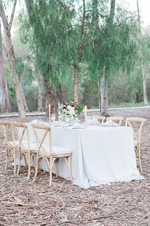 Into the woods outdoor romantic wedding shoot table set up white table linen with light brown wood chairs and white and purple flower centerpiece decor with white place settings and light blue linen napkin decor wedding photo idea