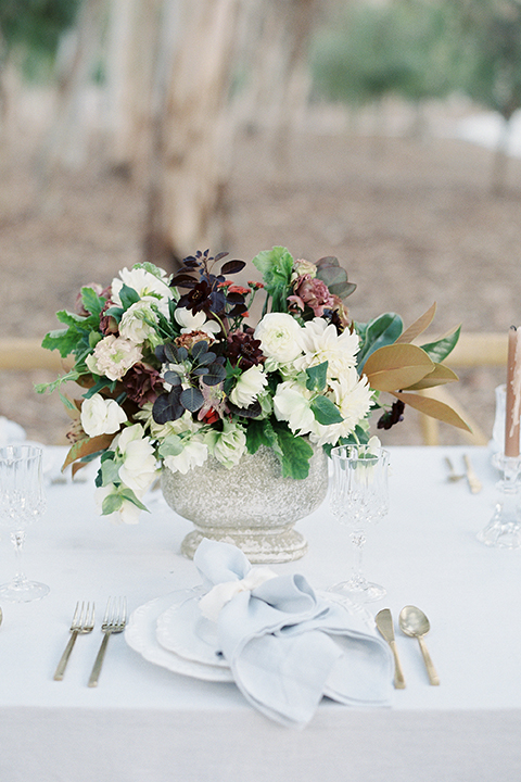 Into the woods outdoor romantic wedding shoot table set up white table linen with light brown wood chairs and white and purple flower centerpiece decor with white place settings and light blue linen napkin decor wedding photo idea