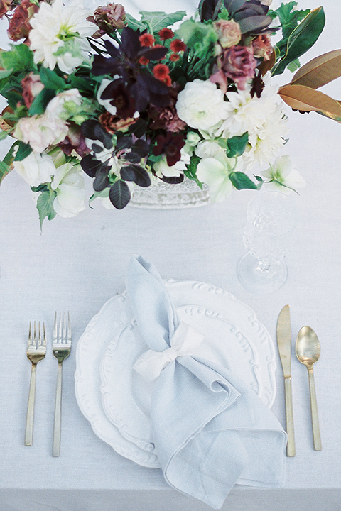 Into the woods outdoor romantic wedding shoot table set up white table linen with light brown wood chairs and white and purple flower centerpiece decor with white place settings and light blue linen napkin decor wedding photo idea