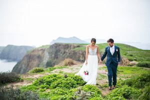 Channel islands wedding shoot bride ball gown with lace bodice and thin straps with plunging neckline and open back design with groom blue suit with matching vest and white dress shirt with long silver striped tie and pink and white floral boutonniere holding hands