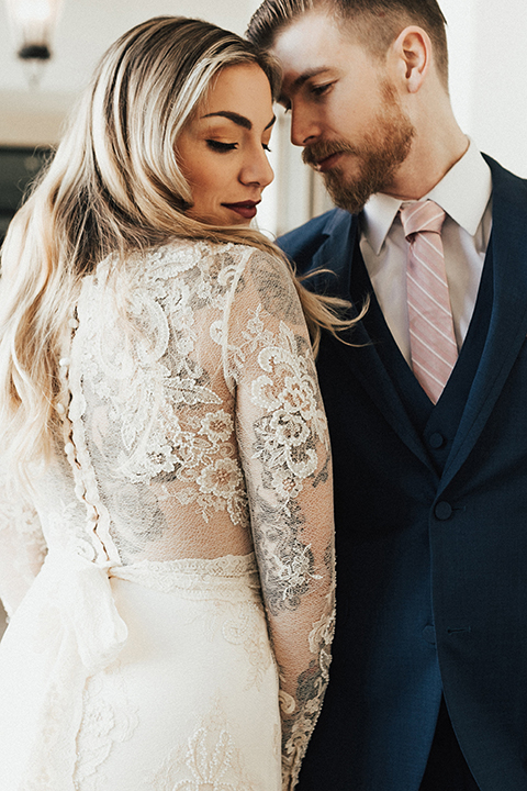 Dana point outdoor wedding shoot at monarch beach resort bride form fitting lace gown with long sleeves and high neckline with groom navy blue suit with matching vest and white dress shirt with long blush pink striped tie and matching pocket square hugging
